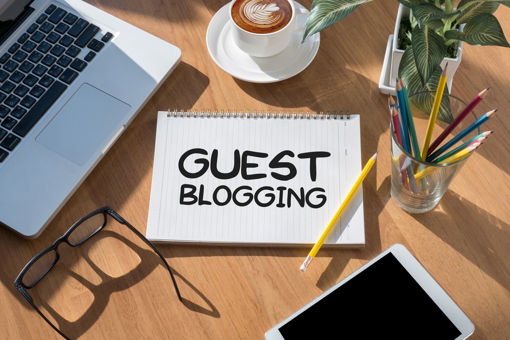 Imagen de un ordenador portátil, una tableta, unas gafas, un lápiz, un café y un bloc de notas con las palabras "guest blogging" escritas.